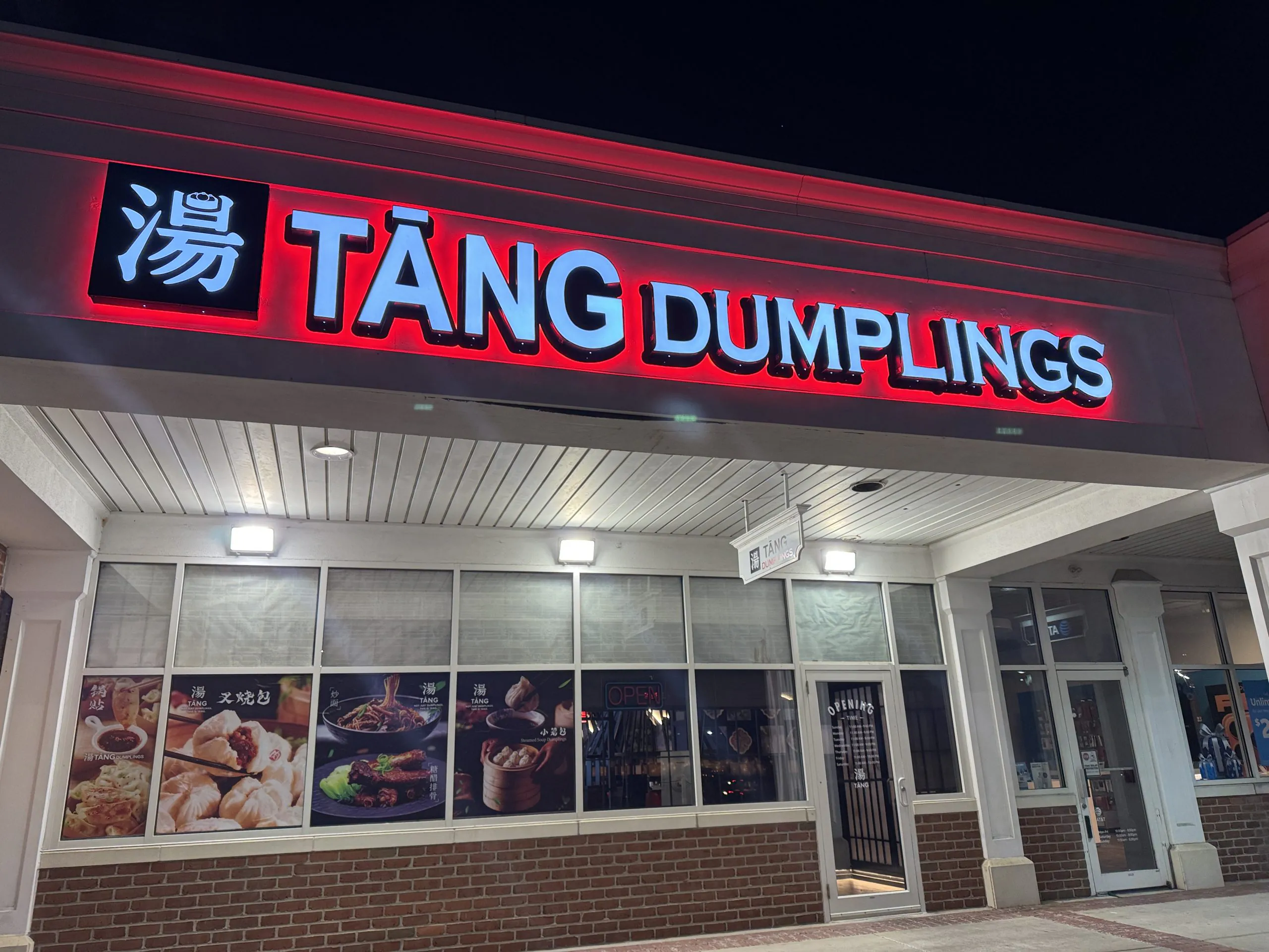 The exterior of Tang Dumplings at night, featuring its bright red and white neon sign above the entrance and menu posters in the windows.